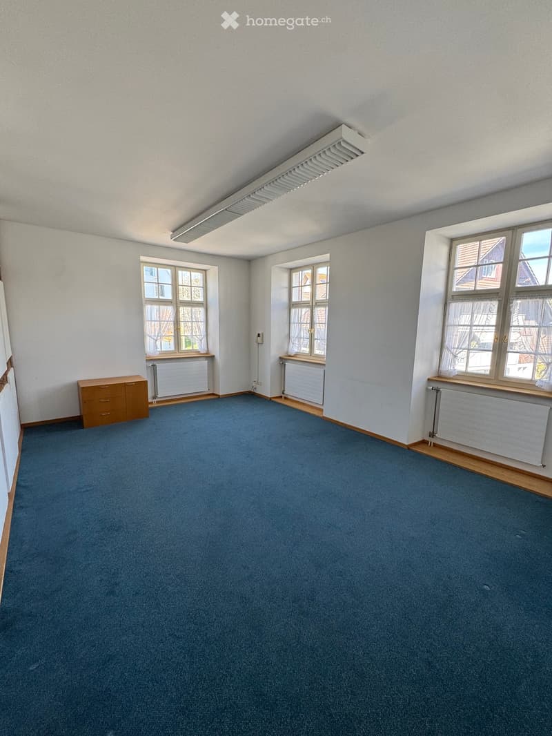 Empty room with blue carpet, white walls, wooden baseboards, and three windows with white frames and curtains. A small wooden cabinet is visible on the left side.
