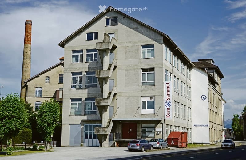 Multi-story industrial-style building with a brick facade, large windows, and a prominent chimney. The building has a modern entrance with a staircase leading up to the main door. Several cars are parked in front of the building.