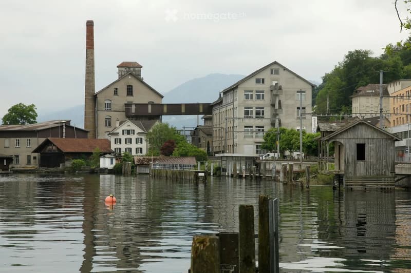 A waterfront view of a historic industrial area with a large brick factory building, a tall chimney, and several smaller structures along the water's edge. The scene includes a calm river, wooden docks, and a small orange buoy floating in the water.