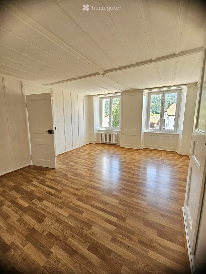 Empty room with wooden flooring, white walls, large windows, and a radiator. No furniture or equipment present.