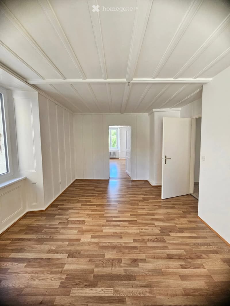 An empty room with wooden flooring, white walls, and a slanted white ceiling. There are two open doors leading to other rooms.