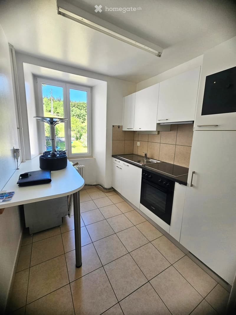 Modern kitchen with white cabinets, stainless steel sink, oven, and a small table with a stool near a large window.