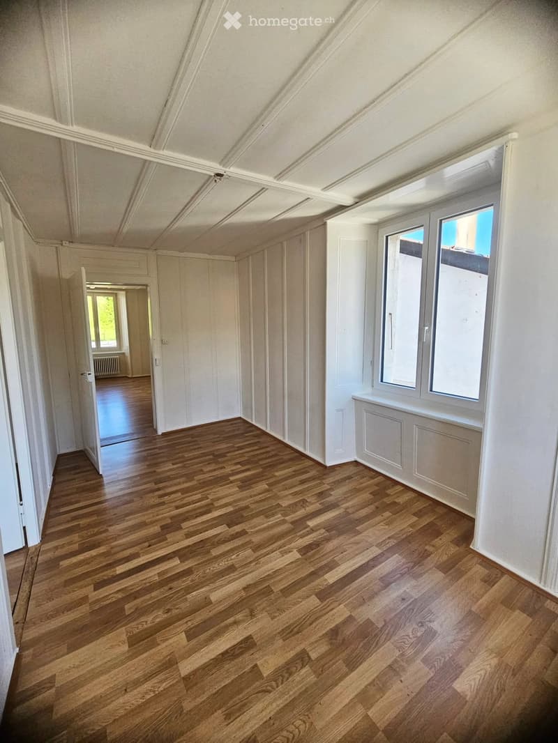Empty room with wooden flooring, white walls, and large windows. The room has a slanted ceiling and an open doorway leading to another room.