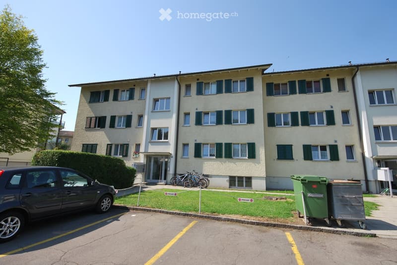 A three-story residential building with light-colored walls and green shutters, featuring multiple windows and a small entrance area with bicycles parked nearby. The building is surrounded by a well-maintained lawn and a paved parking area.