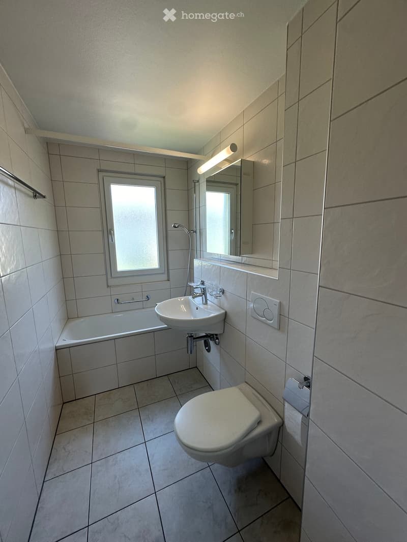 Modern bathroom with tiled walls and floor, featuring a toilet, sink, bathtub, and mirror with built-in lighting.
