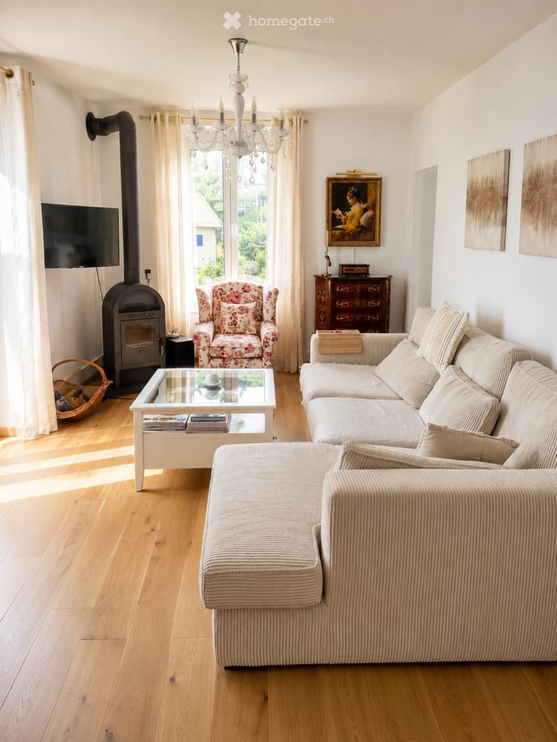 Bright living room with wooden flooring, large window, fireplace, sofa, armchair, coffee table, chandelier, and TV.