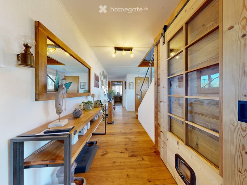 A well-lit hallway with wooden flooring, a wooden shelf on the left side, and a staircase leading upwards. The hallway features modern lighting fixtures and decorative items on the shelf.