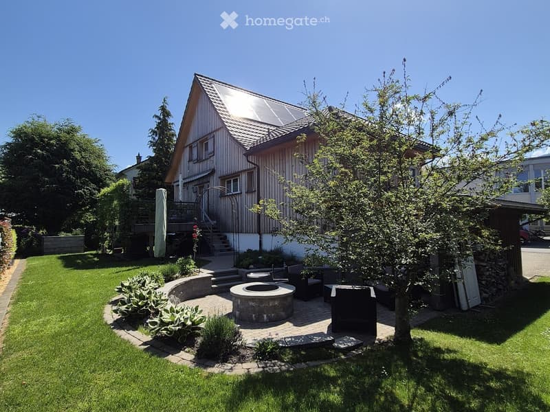 Well-maintained garden with a stone fire pit, seating area, and lush greenery surrounding a modern house with a solar panel roof.