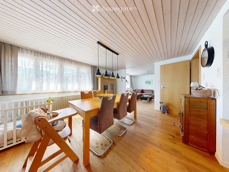 Bright, open-plan living and dining area with wooden floors, a dining table with chairs, a baby chair, a sofa, and a small table lamp on a cabinet.