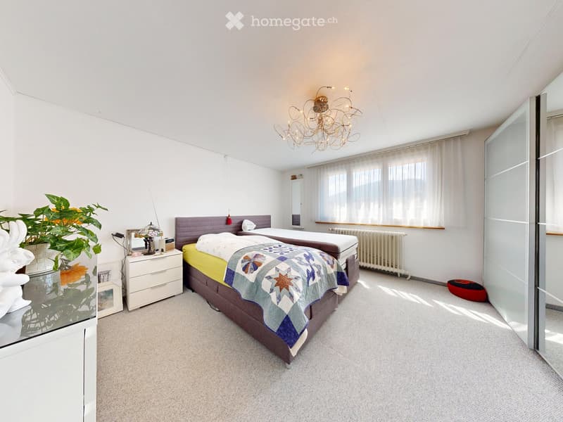 Bright bedroom with two single beds, nightstand, mirror, chandelier, radiator, and plant. The room features large windows with white curtains and a modern glass wardrobe.
