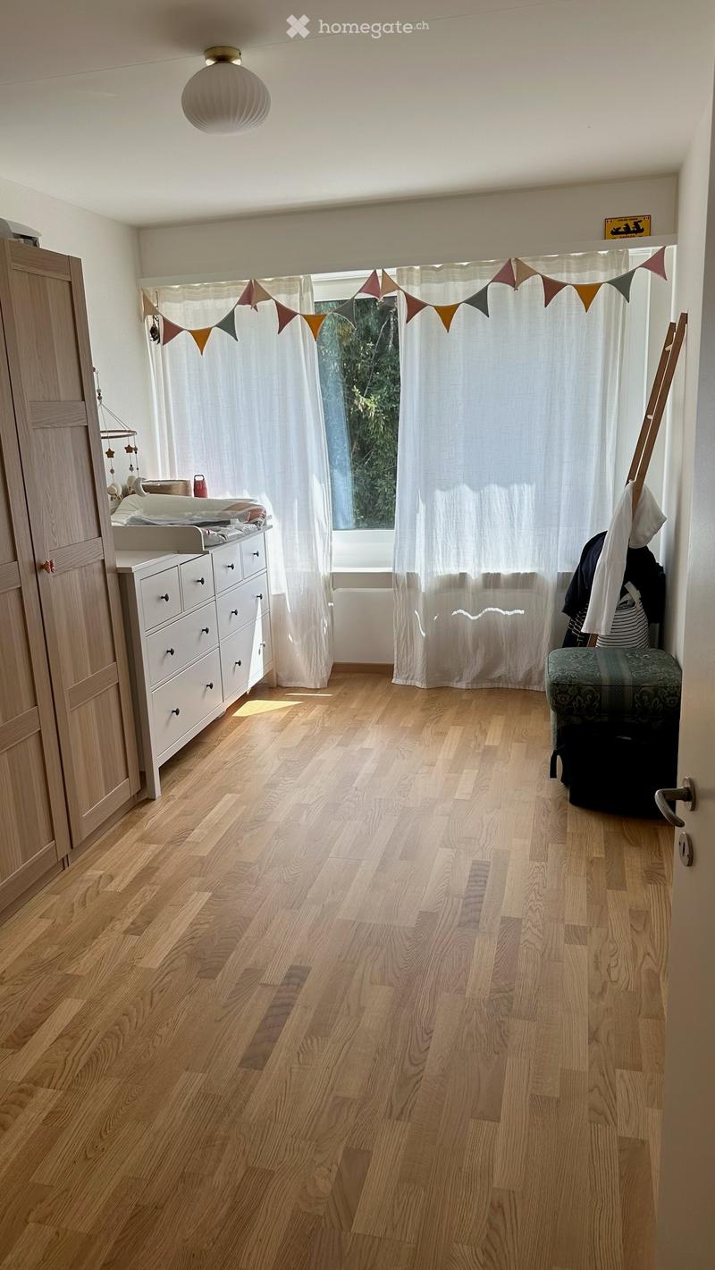 A well-lit room with wooden flooring, a large window with white curtains, a wardrobe, a drawer unit, a small stool, and a decorative banner above the window.