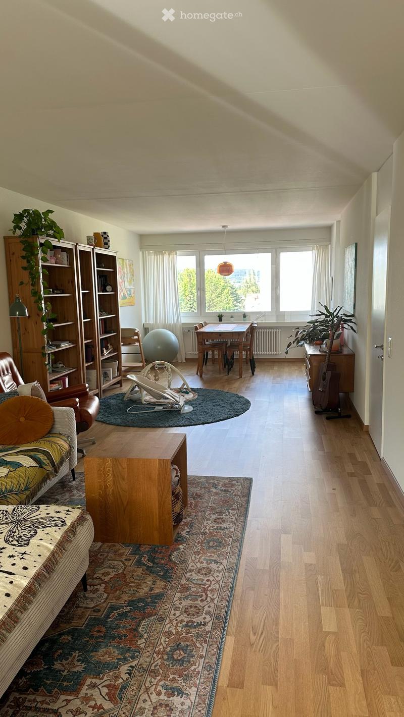 Bright living room with wooden floors, large windows, and a mix of seating and dining areas. Features include a patterned rug, a round coffee table, a sofa, a dining table with chairs, a bookshelf, and a guitar.