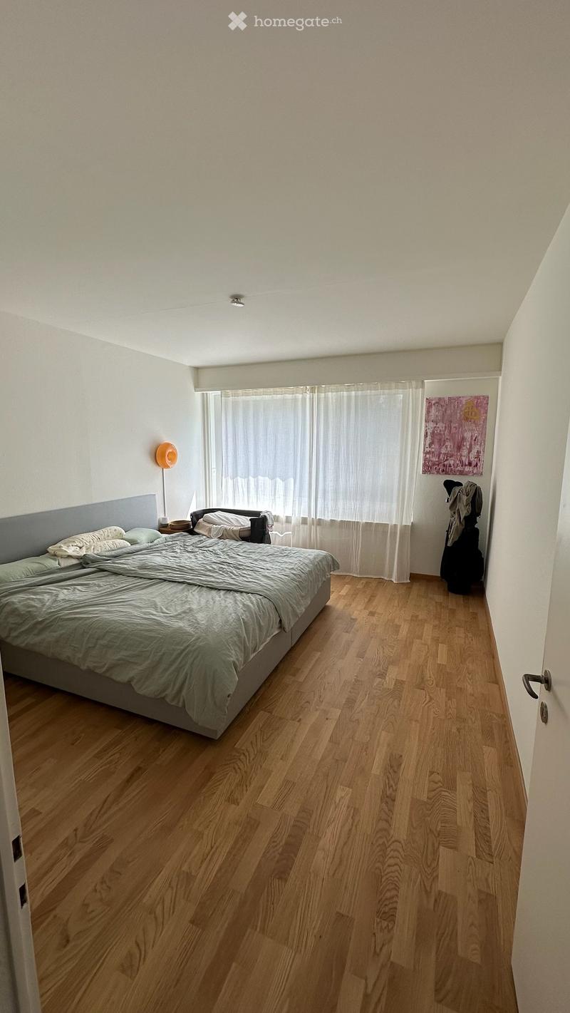 Bedroom with wooden flooring, a large bed with grey bedding, a nightstand with a lamp, a window with white curtains, and a colorful painting on the wall.
