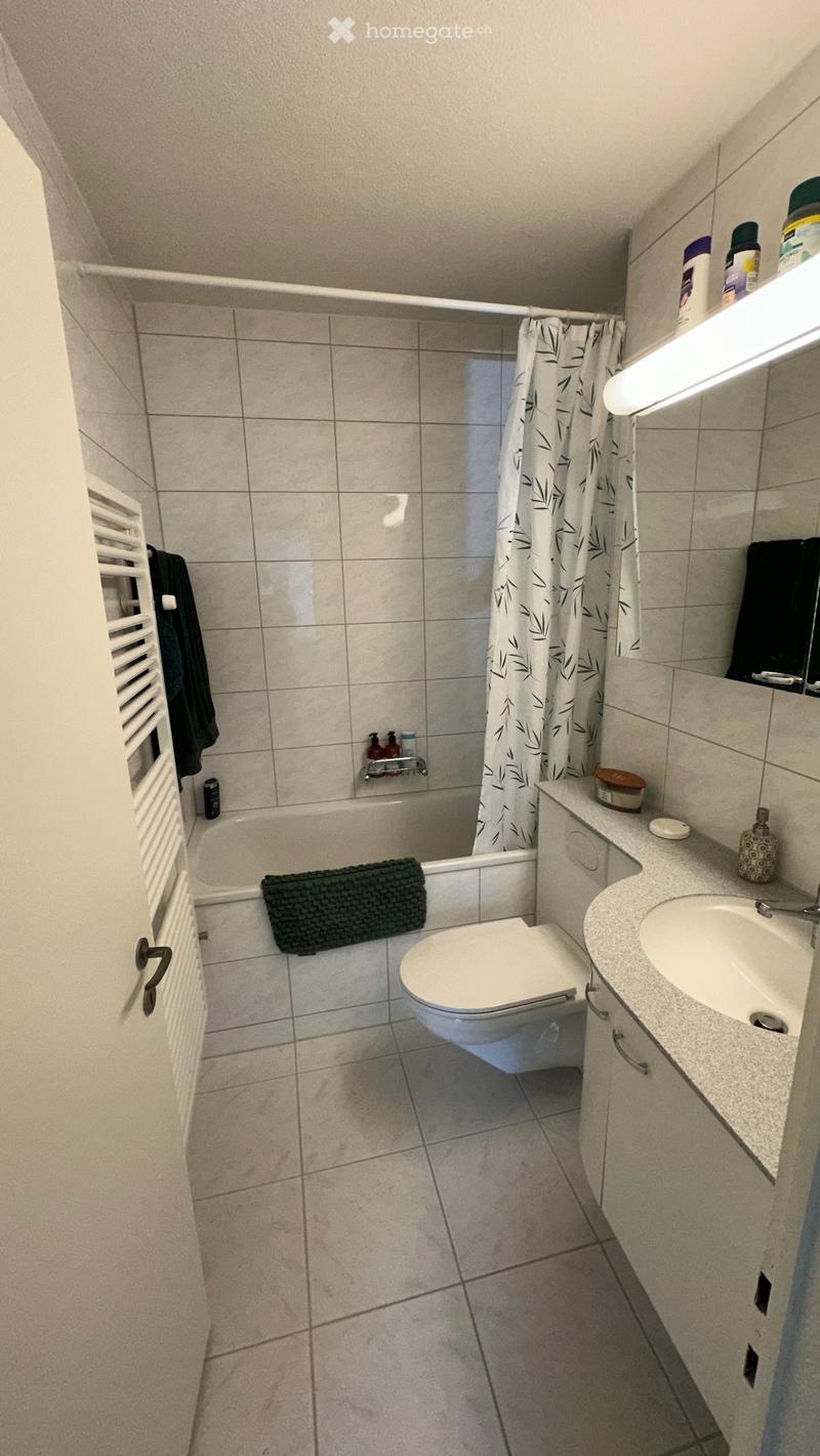 A small bathroom with a toilet, sink, bathtub, and mirror. The bathtub has a green towel on its edge, and the sink has a bottle and a cup on the counter. A shower curtain with a leaf pattern is visible in the bathtub area.