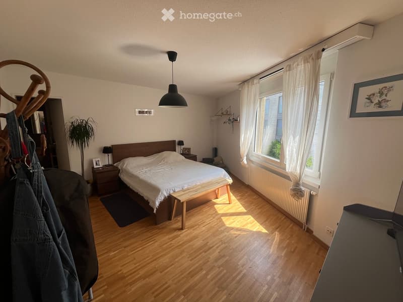 Bright bedroom with wooden flooring, large bed with white bedding, nightstand, bench at the foot of the bed, and large windows with white curtains.
