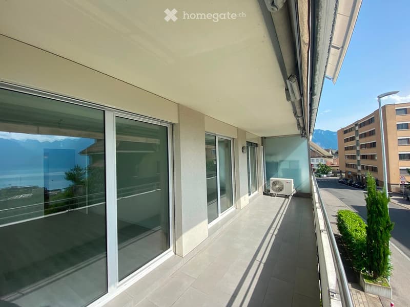 Balcony with large glass sliding doors, tiled floor, metal railing, and an air conditioning unit. The view shows a street, buildings, and mountains in the distance.