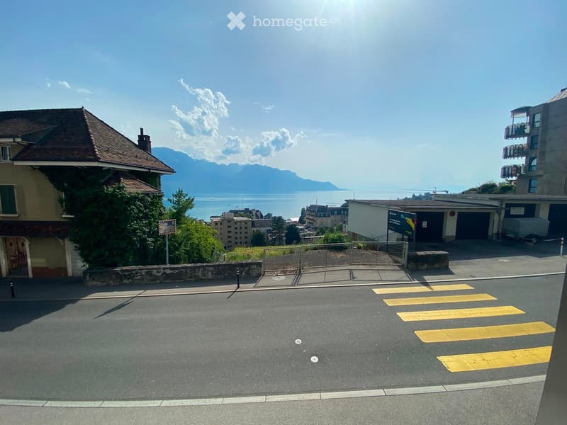 View of a parking area with yellow crosswalk markings, surrounded by buildings and a scenic view of a lake and mountains in the background.