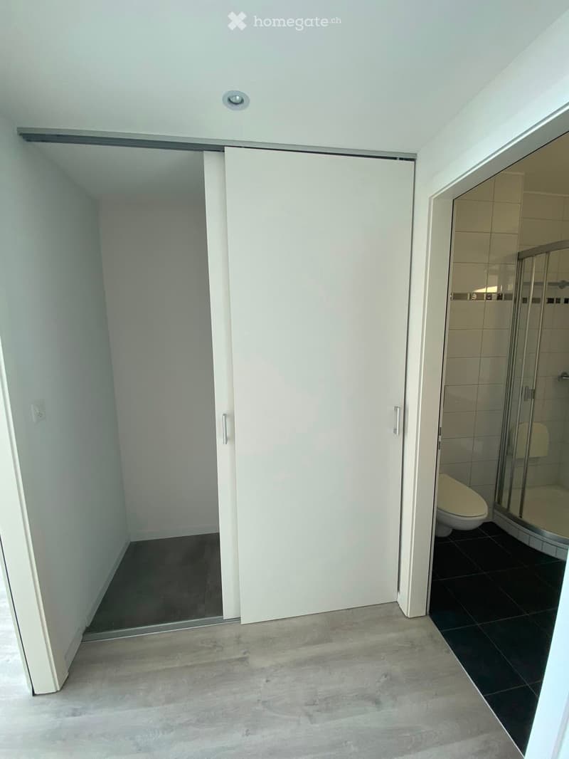 A narrow hallway with light-colored walls and a gray tiled floor, leading to a bathroom with a toilet and a glass-enclosed shower stall.