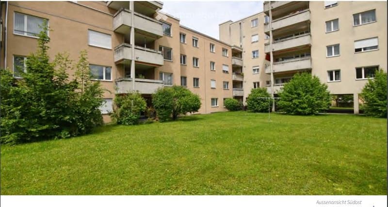A grassy courtyard surrounded by multi-story apartment buildings with balconies and windows. The area is well-maintained with trimmed bushes and trees.