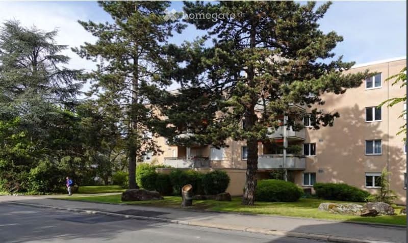Multi-story residential building with balconies, surrounded by trees and greenery. The building has a light beige facade and multiple windows.