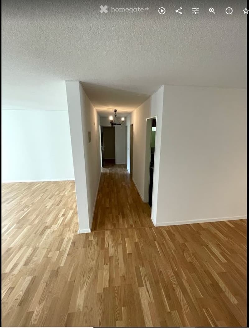 Empty hallway with wooden flooring and white walls, featuring a chandelier at the far end.