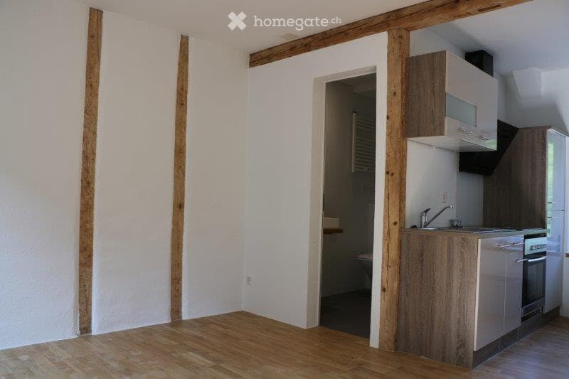 Modern kitchen with wooden flooring, white walls, wooden beams, a sink, faucet, cabinet, and built-in appliances like a microwave, oven, and refrigerator.