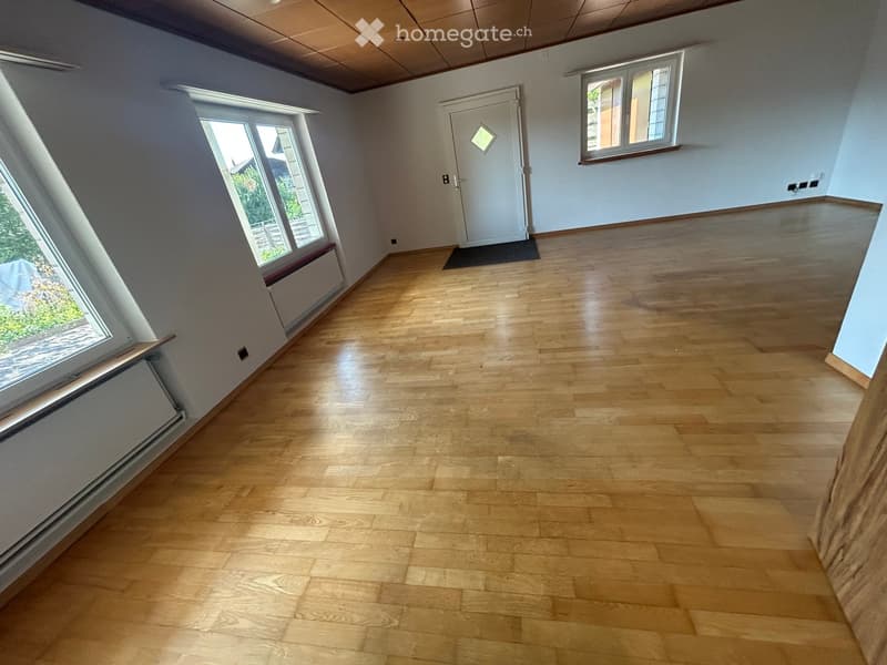 Empty room with wooden flooring, light blue walls, and large windows. The space is unfurnished with a door leading to another area.