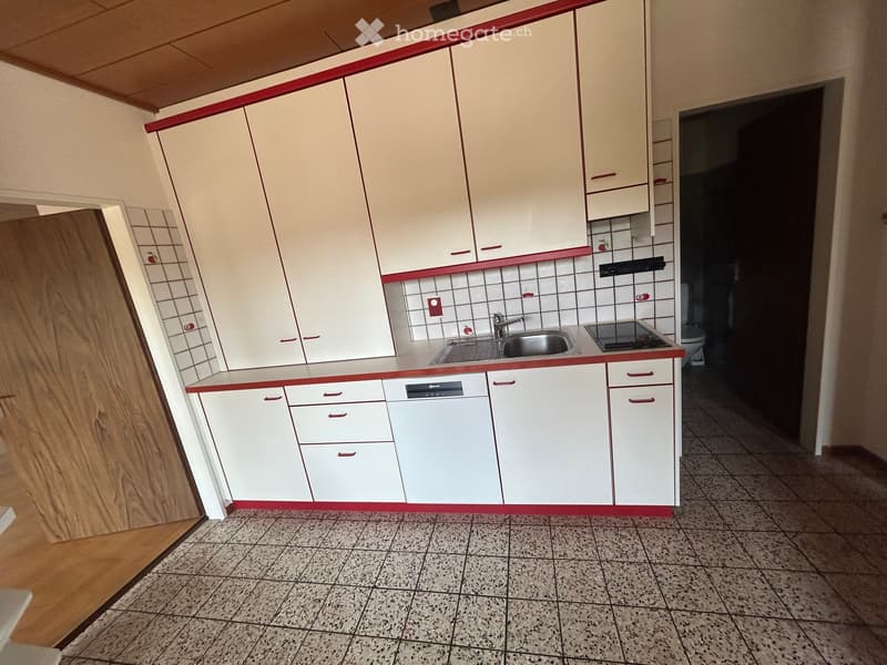 Modern kitchen with white cabinets, red trim, a sink, a stove, and a dishwasher. The floor is tiled, and there is a visible bathroom area in the background.