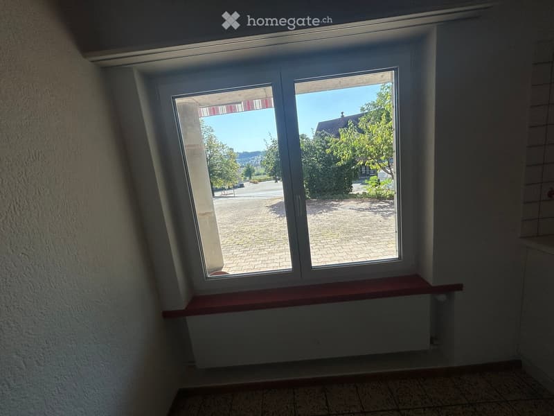 Empty room with large windows offering a view of a paved outdoor area and greenery. Features a red window sill and tiled flooring.