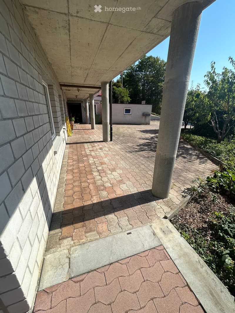 Brick-paved covered walkway with concrete columns and walls, leading to an open area with greenery and a small building.