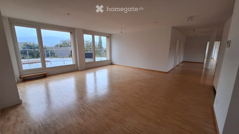 Empty room with wooden flooring, large windows, and a small bench near the window. No furniture or equipment present.