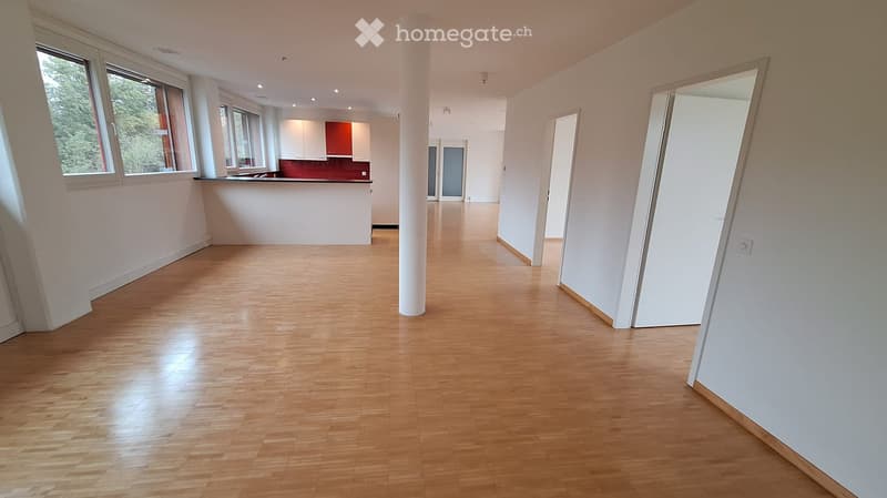 Empty room with wooden flooring, white walls, large windows, and a visible kitchen area in the background.