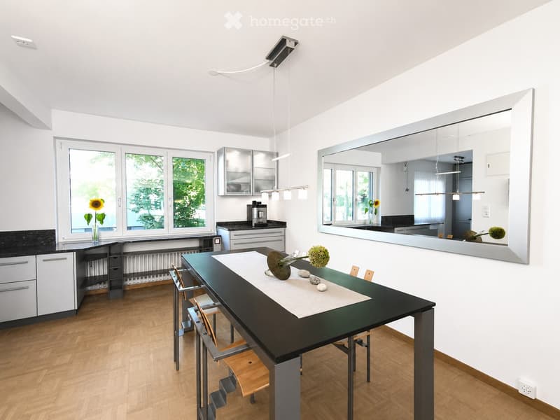 Modern kitchen with white cabinets, black countertops, a large dining table with chairs, and a coffee machine. The room features large windows with a view of greenery and a decorative mirror on the wall.