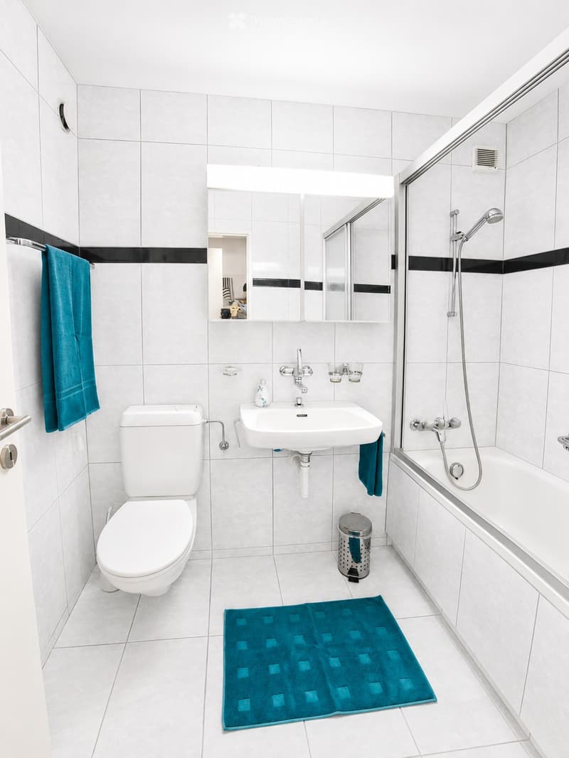Modern bathroom with white tiles, a toilet, a sink with a mirror above it, a bathtub with a showerhead, and a blue bathmat on the floor.