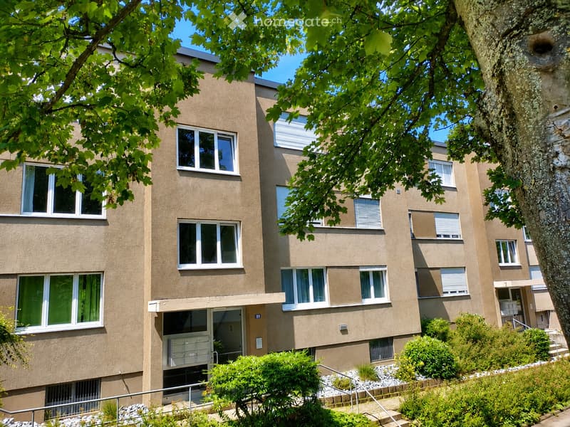 Multi-story residential building with beige facade, multiple windows, and a small entrance area surrounded by greenery.