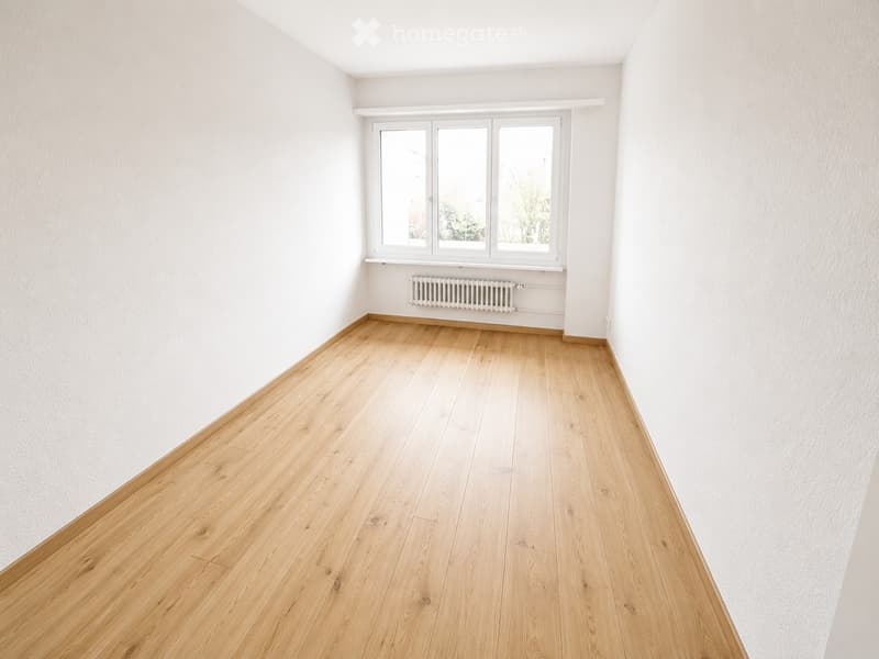 Empty room with light wooden flooring and white walls, featuring a window with white frames and a radiator below it.