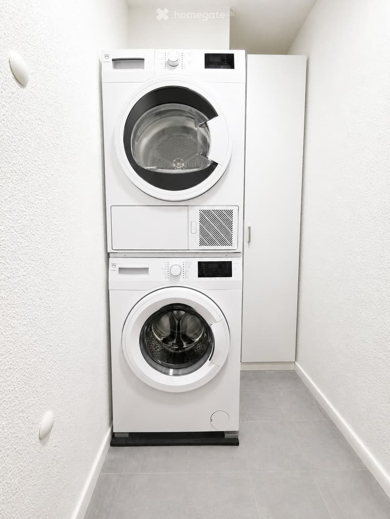 A small bathroom with a stacked washing machine and dryer unit, white walls, and grey tiled flooring.