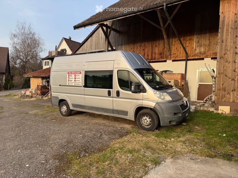A silver van with a sticker saying 'Die Aikrounder' parked in front of a building with wooden walls and a red roof. The van is parked on a gravel driveway.