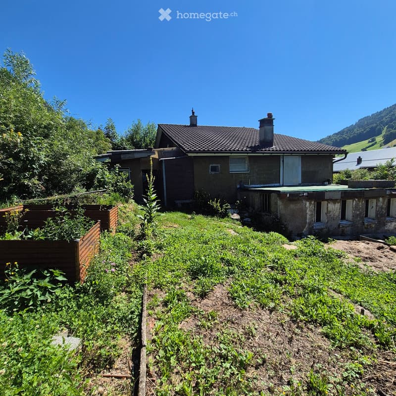 A two-story house with a tiled roof and chimneys, surrounded by greenery and a garden area with plants and a wooden fence.