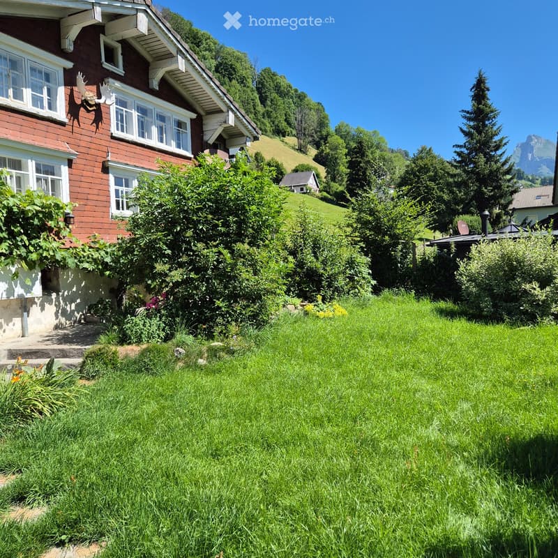 A well-maintained garden with green grass, bushes, and trees, leading up to a two-story house with red brick walls and white-framed windows. The house is surrounded by lush greenery and a scenic mountain backdrop.