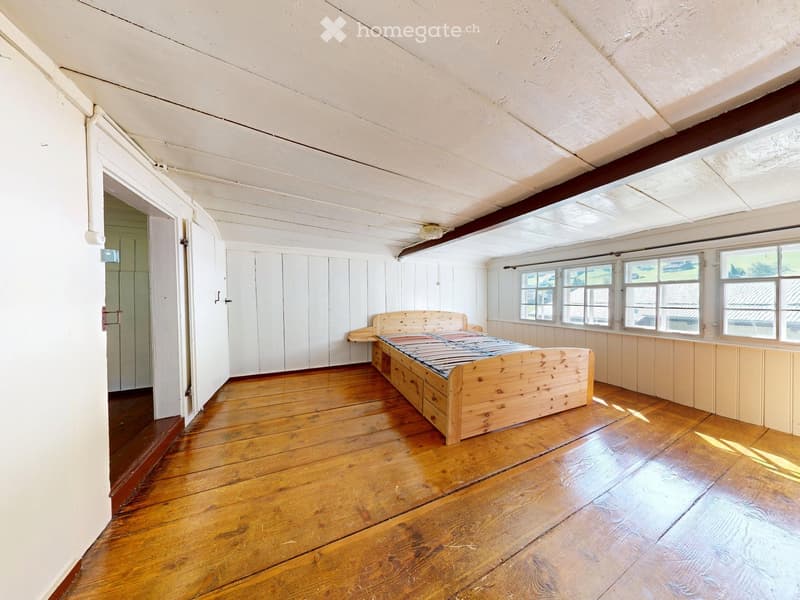 Spacious bedroom with wooden floor, white paneled walls, large windows, and a simple wooden bed.