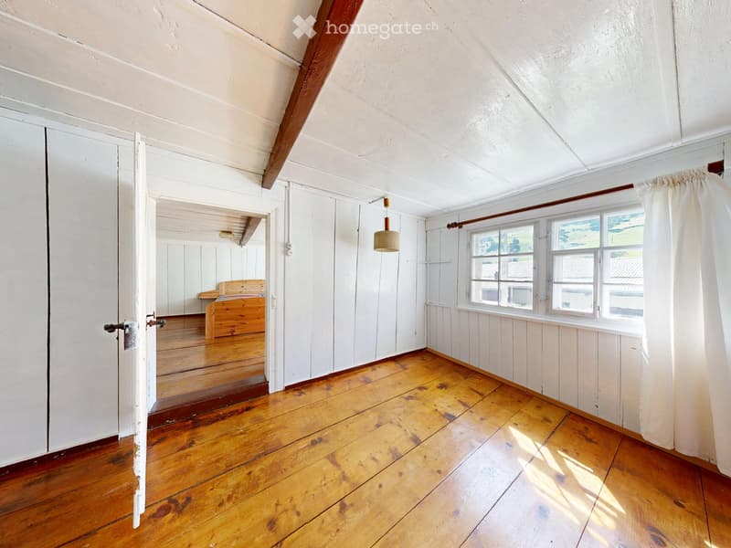 Empty room with wooden flooring, white walls, and exposed wooden beams on the ceiling. Features a large window with white curtains and an open doorway leading to another room.