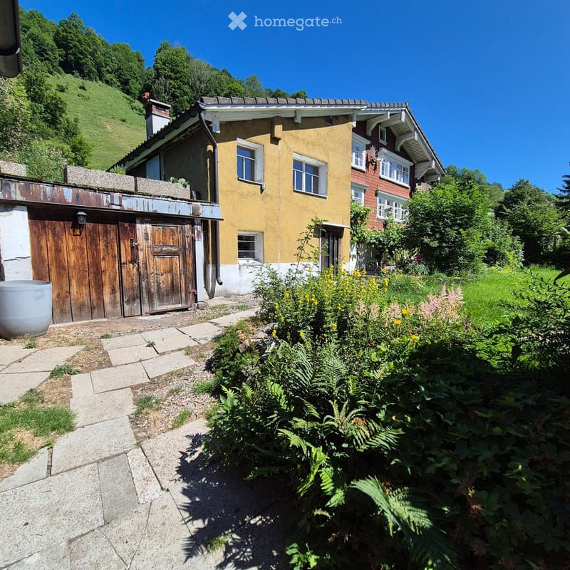 Two-story house with yellow and red exterior, wooden garage, garden with flowers, and a raised ground floor.