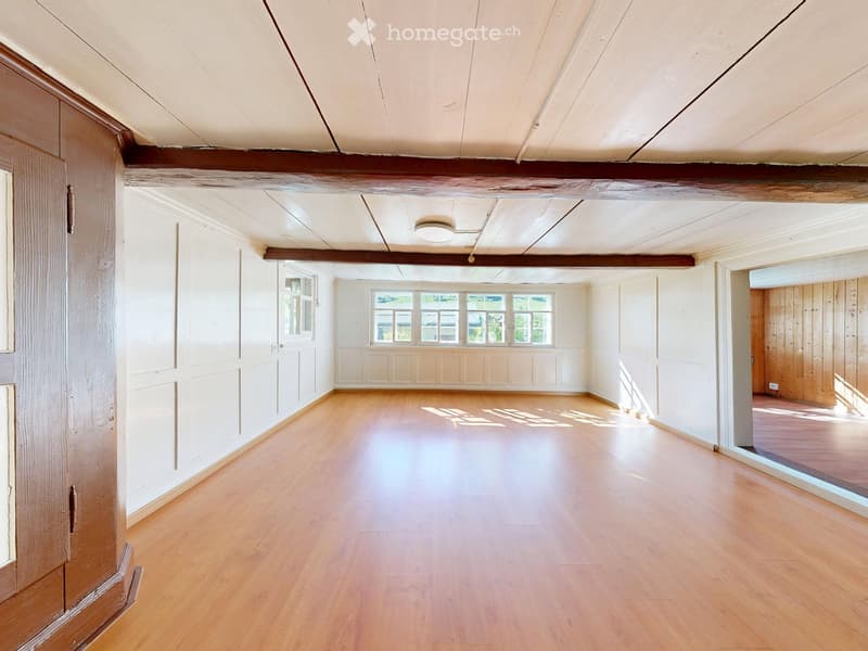 Empty room with wooden flooring, white walls, and large windows allowing natural light. Exposed wooden beams on the ceiling.