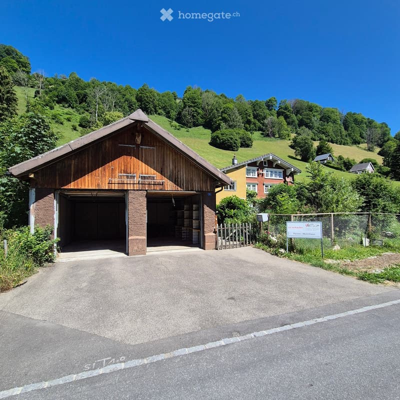 A large wooden garage with a sloped roof and stone base, situated on a paved driveway with a fenced area and a sign nearby.