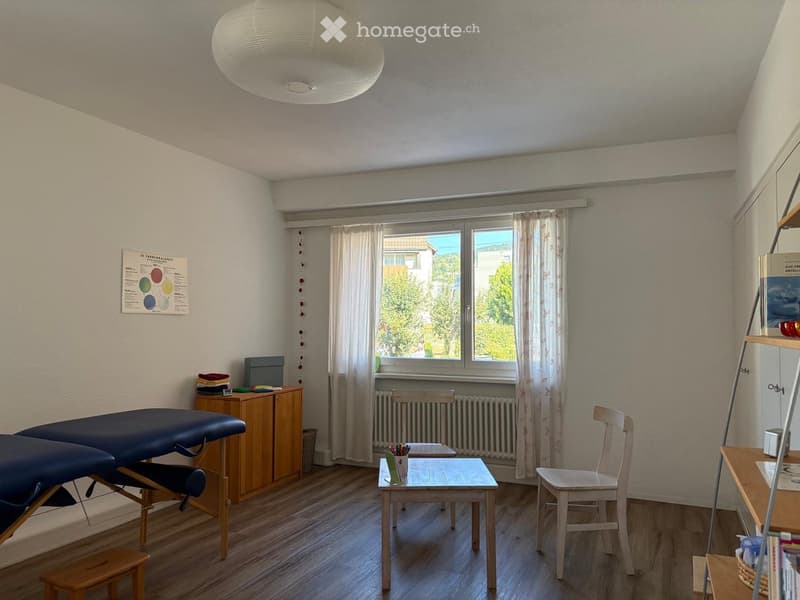 A well-lit room with wooden flooring, a massage table, a small table with chairs, a wooden cabinet, and a bookshelf. The room has large windows with curtains and a poster on the wall.