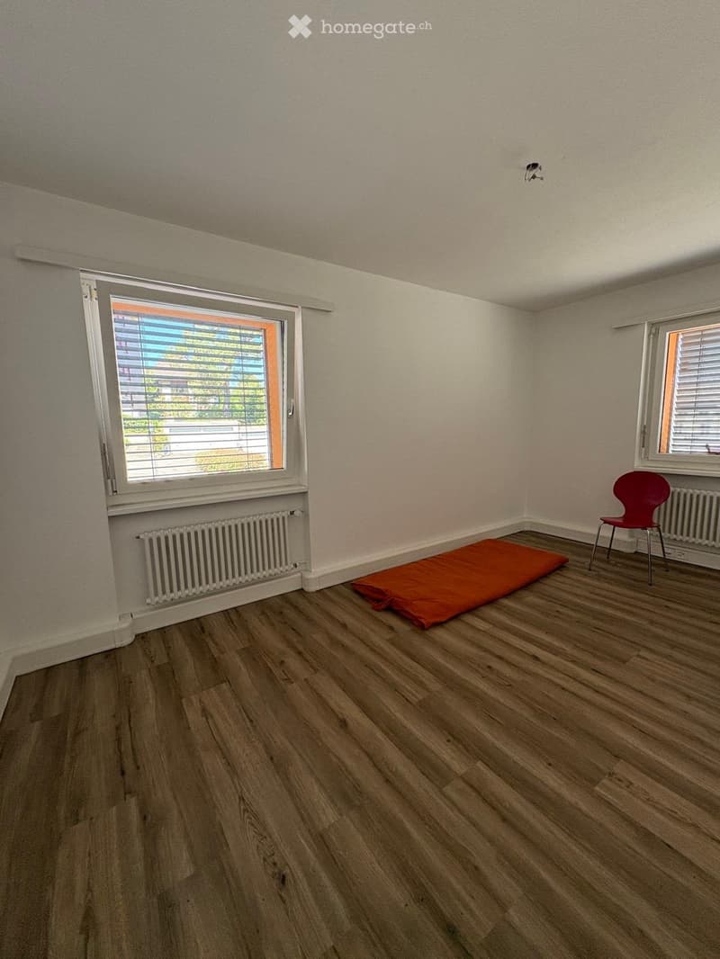 A small, unfurnished room with wooden flooring, two windows with blinds, radiators, and a single red chair.