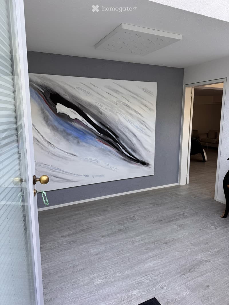 Empty room with grey walls, light wooden flooring, and a large abstract painting on one wall. A glass door with blinds is visible on the left, and an open doorway leads to another room with furniture visible in the background.