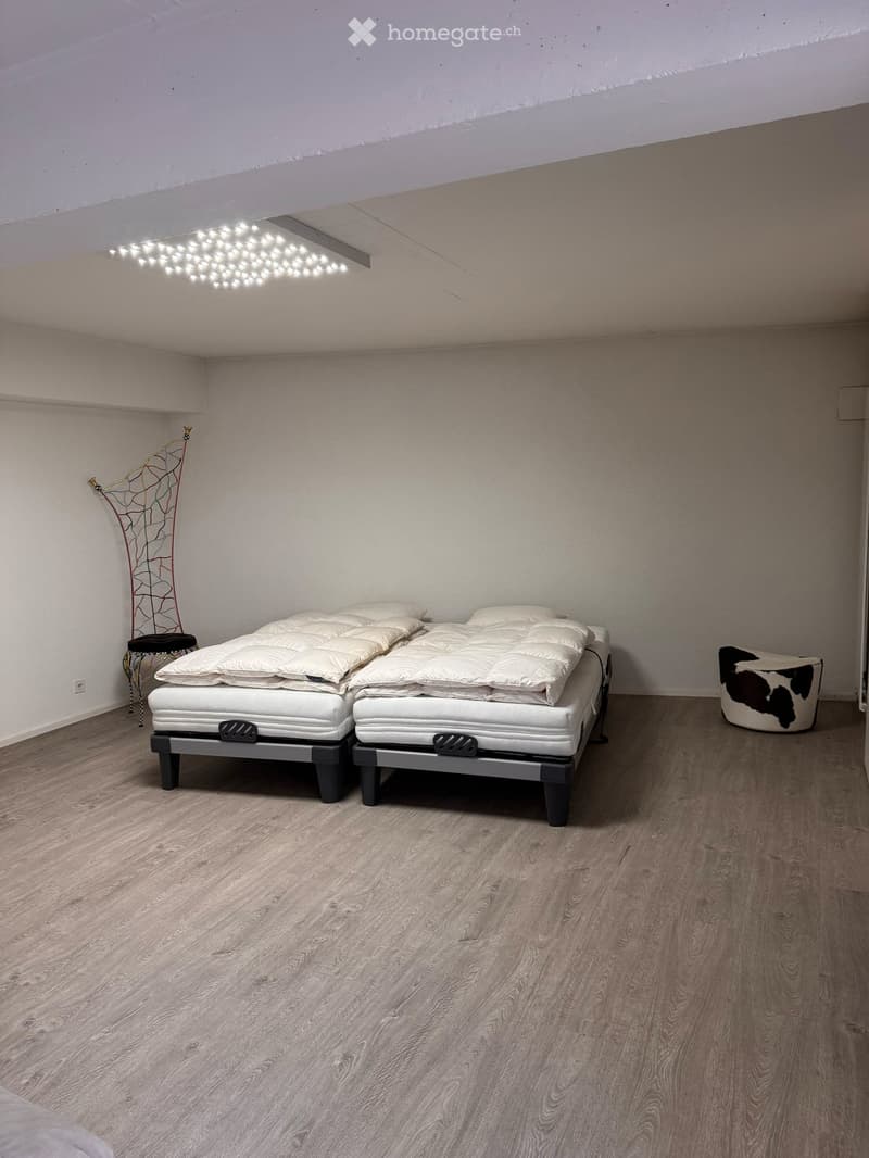 Modern bedroom with two single beds, a decorative chair, and a small stool, featuring light wood flooring and white walls.