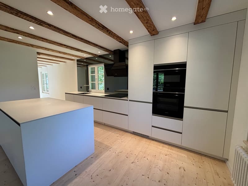 Modern kitchen with white cabinetry, a central island, and integrated appliances including an oven and range hood. Exposed wooden beams on the ceiling and wooden flooring.